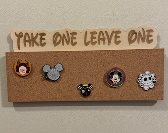 May include: A wooden sign with the text "TAKE ONE LEAVE ONE" above a corkboard. The corkboard displays five decorative pins with various designs, including a Mickey Mouse head and a skull.