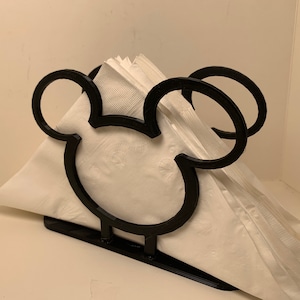 May include: Black metal napkin holder shaped like Mickey Mouse's head. The holder is empty and ready to hold napkins.