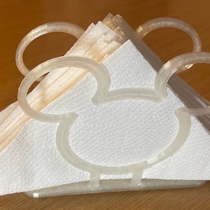May include: A white plastic napkin holder shaped like Mickey Mouse's head. The holder has two large, rounded ears and a small, rounded nose.