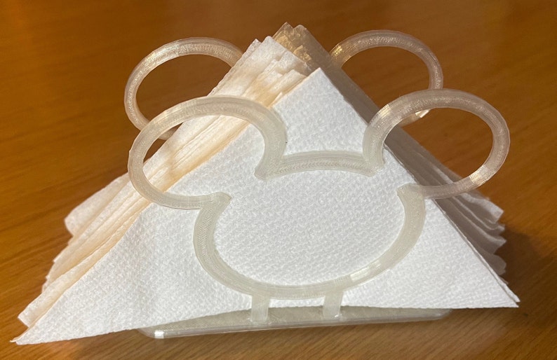 May include: A white plastic napkin holder shaped like Mickey Mouse's head. The holder has two large, rounded ears and a small, rounded nose.