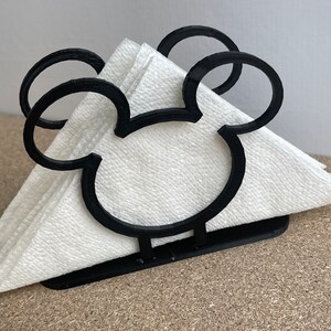 May include: Black napkin holder shaped like Mickey Mouse's head. The holder is made of plastic and has a white napkin inside.
