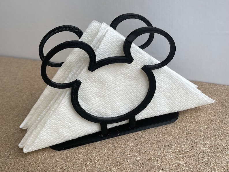 May include: Black napkin holder shaped like Mickey Mouse's head. The holder is made of plastic and has a white napkin inside.