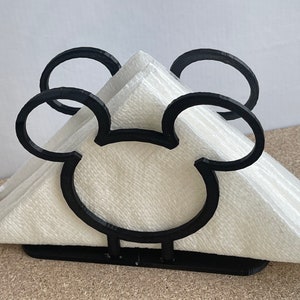 May include: Black napkin holder shaped like Mickey Mouse's head. The holder is made of plastic and has a white napkin inside.