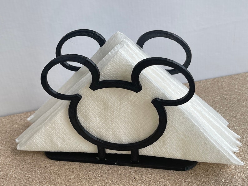 May include: Black napkin holder shaped like Mickey Mouse's head. The holder is made of plastic and has a white napkin inside.