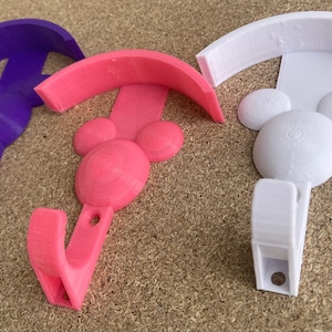 May include: Three 3D printed wall hooks in the shape of Mickey Mouse heads. The hooks are in purple, pink, and white.