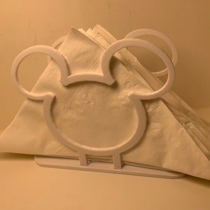 May include: White plastic napkin holder shaped like Mickey Mouse's head. The holder has two large ears and a small base.