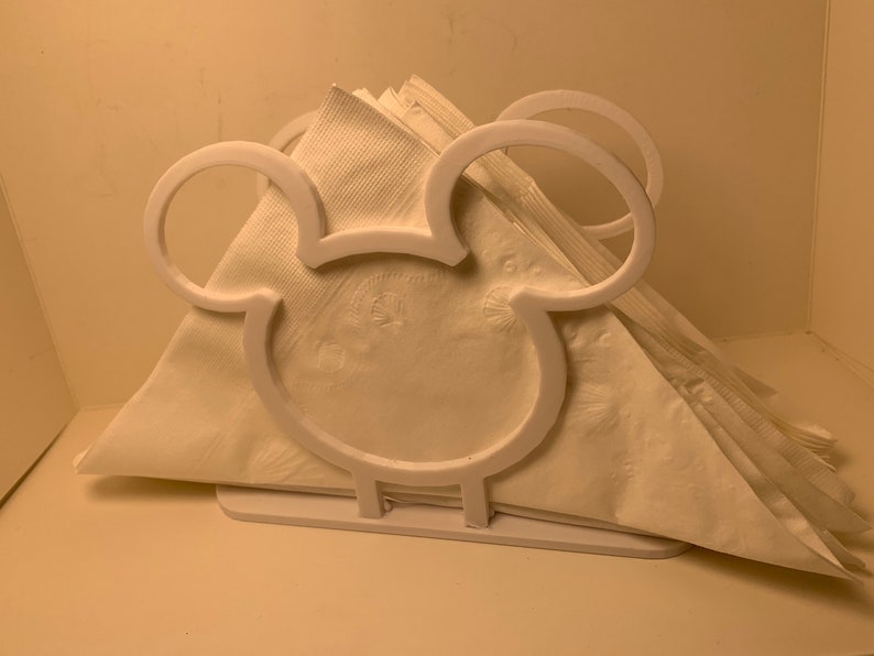 May include: White plastic napkin holder shaped like Mickey Mouse's head. The holder has two large ears and a small base.