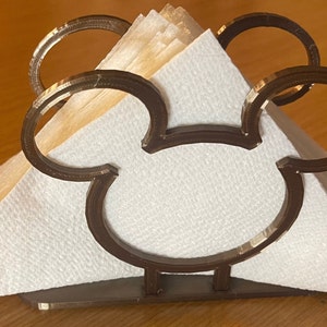 May include: A brown metal napkin holder shaped like Mickey Mouse's head. The holder has two large circles for the ears and a smaller circle for the face. The holder is holding a stack of white napkins.