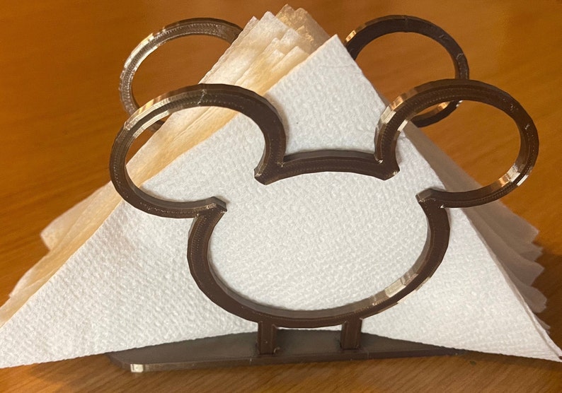 May include: A brown metal napkin holder shaped like Mickey Mouse's head. The holder has two large circles for the ears and a smaller circle for the face. The holder is holding a stack of white napkins.