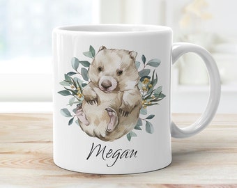 Op de afbeelding: Witte keramische mok met een aquarel-illustratie van een wombat omringd door groene bladeren en gele bloemen. De naam "Megan" staat in cursief onderaan de mok.