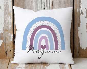 Pode incluir: Travesseiro branco com um design de arco-íris em tons de roxo, azul e cinza. O arco-íris tem bolinhas brancas e um pequeno coração no centro. O nome "Megan" está escrito em itálico abaixo do arco-íris.