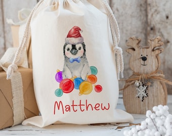 May include: A white cotton gift bag with a penguin wearing a Santa hat and a blue bow tie surrounded by colorful ornaments. The bag is personalized with the name "Matthew".