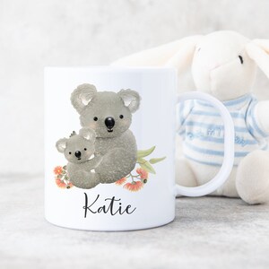 May include: White ceramic mug with a design of two koalas, one holding the other, with eucalyptus leaves and flowers. The mug has the name "Katie" printed on the bottom.