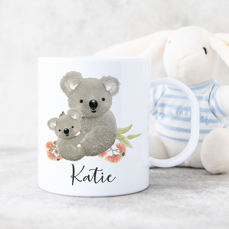 May include: White ceramic mug with a design of two koalas, one holding the other, with eucalyptus leaves and flowers. The mug has the name "Katie" printed on the bottom.