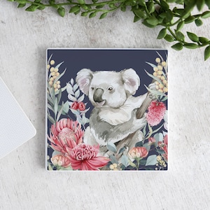 Op de afbeelding: Vierkante onderzetter met een aquarel illustratie van een koala op een tak, omringd door roze en gele bloemen en groen blad tegen een marineblauwe achtergrond. De onderzetter is wit.