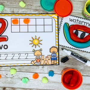 Może przedstawiać: Drukowana karta pracy do nauki liczby dwa. Karta pracy zawiera czerwoną liczbę dwa z plasteliny, słowo "dwa" i linię do śledzenia liczby. Na karcie pracy znajdują się r&oacute;wnież dwa pompony pomarańczowe.