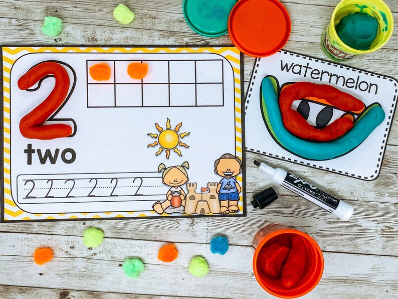 Może przedstawiać: Drukowana karta pracy do nauki liczby dwa. Karta pracy zawiera czerwoną liczbę dwa z plasteliny, słowo "dwa" i linię do śledzenia liczby. Na karcie pracy znajdują się r&oacute;wnież dwa pompony pomarańczowe.