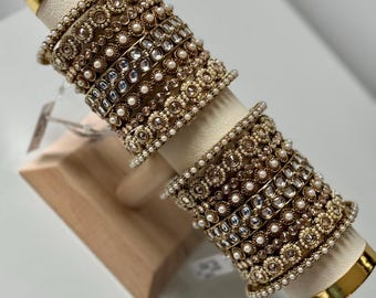 May include: A collection of gold-toned bangles with intricate floral and geometric designs, adorned with small pearls and clear gemstones. The bangles are displayed on a wooden stand with gold accents, showcasing their ornate details.