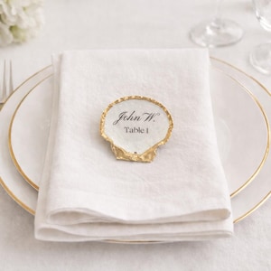 May include: A formal table setting with a white linen napkin, gold-rimmed plates, and silverware. A seashell-shaped place card with the text "John W. Table 1" sits atop the napkin. White flowers are in the background.