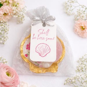 Puede incluir: Un posavasos en forma de concha con borde dorado, dentro de una bolsa transparente plateada. El posavasos tiene un diseño floral. Una etiqueta blanca con el texto "Shell be here soon!" y un gráfico de concha.