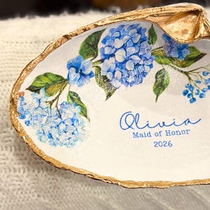 Puede incluir: Un plato de concha blanca con borde dorado, decorado con flores de hortensia azules y hojas verdes. El plato está personalizado con el nombre "Olivia", "Dama de honor" y el año "2026" en escritura azul.
