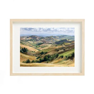 May include: A framed landscape painting depicting rolling hills in shades of green, gold, and brown, under a cloudy sky. The artwork is presented in a light wood frame. The painting captures a serene countryside scene.