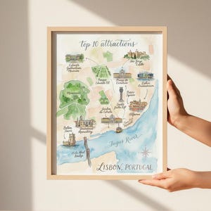 May include: Watercolor map print of Lisbon, Portugal, featuring the top 10 attractions. The map includes illustrations of landmarks like Jerónimos Monastery, Belém Tower, and São Jorge Castle, with the Tagus River in blue. The print is in a light wooden frame.