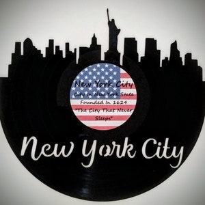 May include: A black vinyl record with a white silhouette of the New York City skyline, including the Statue of Liberty. The record has a sticker with an American flag and the text "New York City, City in New York State, Founded in 1624, "The City That Never Sleeps."