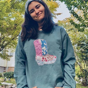 Könnte beinhalten: Türkisfarbenes Sweatshirt mit einem Patchwork-Cowboy-Stiefel-Design. Der Stiefel besteht aus floralem und gemustertem Stoff in Rosa, Blau und Weiß. Die Person trägt ein gemustertes Stirnband und Jeans.