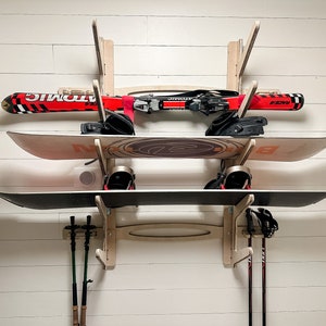 May include: A wooden wall-mounted ski and snowboard rack holding a pair of skis, two snowboards, and ski poles. The skis are red and black, and the snowboards are white and black. The rack is mounted on a white wall.