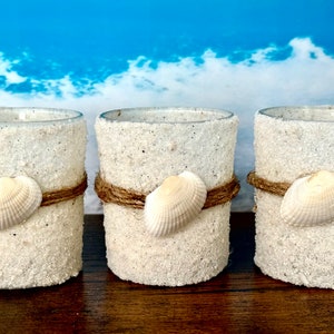 May include: Three glass candle holders with a white sand-like texture and a seashell glued to the front. Each holder is wrapped with a brown twine.