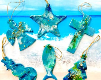 Op de afbeelding: Zes hangende ornamenten in verschillende vormen, waaronder een engel, een ster, een kruis, een sneeuwpop, een vis en een kerstboom. Elk ornament is gemaakt van blauwe en groene hars met ingebed zand en kleine schelpfragmenten, voorzien van touw.
