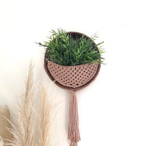 May include: A circular macrame wall planter in brown and beige tones, filled with green artificial foliage. The planter features a woven design and a long tassel in a matching colour. The wall decor is mounted on a white wall, with dried pampas grass in the foreground.