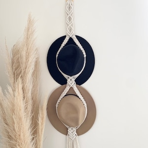 Puede incluir: Un perchero de macramé blanco con dos sombreros, uno negro y otro marrón, colgados en él. El perchero está sujeto a una pared blanca con un anillo de madera.