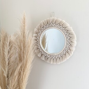 May include: A circular macrame mirror in beige, with a fringed edge, hanging on a white wall. The mirror's frame is made of woven rope and has a circular design with a fringe of knotted threads. Pampas grass is in the foreground.
