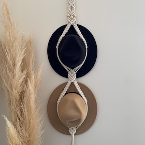 May include: A white macrame hat rack with wooden beads hangs on a white wall. Two hats are displayed on the rack, one black and one brown.