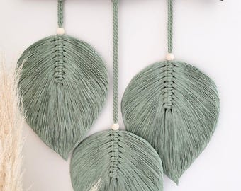 May include: A wall hanging featuring three sage green macrame leaves suspended from a natural wooden branch. Each leaf is crafted with intricate knotted cord, finished with a single light-colored wooden bead at the top, and hangs by a braided cord.