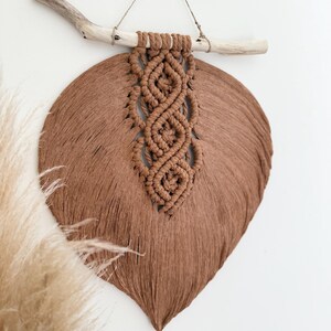 May include: A brown macrame wall hanging shaped like a large leaf, featuring a central column of intricate knotted patterns and fringed edges. The piece is suspended from a light-coloured wooden branch by a thin string attached to the top.