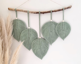 May include: A wall hanging featuring five sage green macrame leaves suspended from a natural wooden branch. Each leaf is crafted from cotton cord with a central knot pattern and a small wooden bead accent, attached to the branch by braided cords.