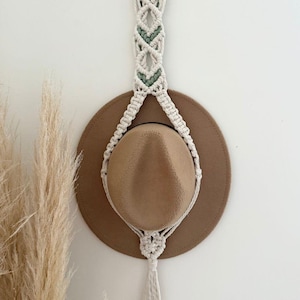May include: A beige macrame hat holder with a wooden ring and a tassel, holding a brown hat. The macrame design includes green accents. The hat holder is hanging on a white wall.