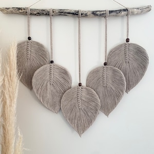 May include: A wall hanging featuring five macrame feathers in a light beige shade. The feathers are suspended from a driftwood branch and arranged in a staggered formation.