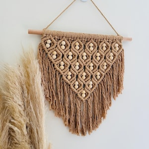 May include: A tan macrame wall hanging featuring a diamond-shaped pattern with integrated light-coloured wooden beads. The piece is suspended from a horizontal wooden dowel by a cord and finished with long, loose fringe at the bottom edge.