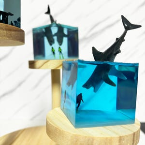 May include: Three clear resin cubes with wooden bases, each containing a different shark figurine. The top cube features a black shark with a diver figurine, the middle cube features a black shark with a diver figurine, and the bottom cube features a white shark with a diver figurine.