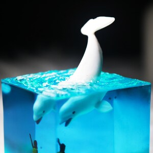 May include: A white whale figurine is submerged in a clear blue resin cube. The whale is leaping out of the water, with its tail in the air. The resin cube is filled with small bubbles, giving the appearance of water.