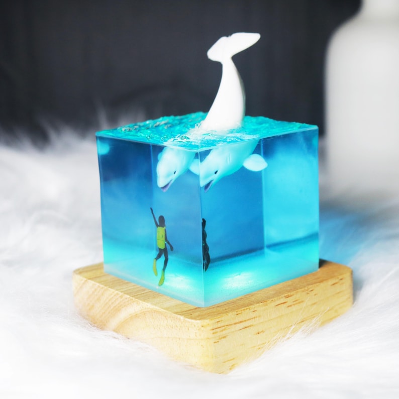 May include: A clear cube filled with blue resin, containing a white whale figurine and a miniature diver figurine. The cube is sitting on a wooden base.