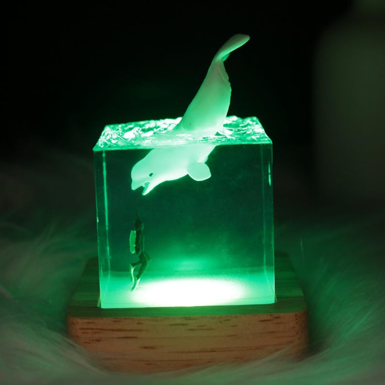 May include: A white whale figurine is suspended in a clear cube filled with green resin, creating a 3D ocean scene. A smaller figurine of a diver is positioned below the whale. The cube sits on a wooden base with a light illuminating the scene from below.