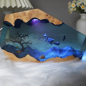 May include: A wooden base with a clear resin insert featuring a witch flying on a broomstick, a tree, and a pumpkin. The resin is illuminated with blue light.