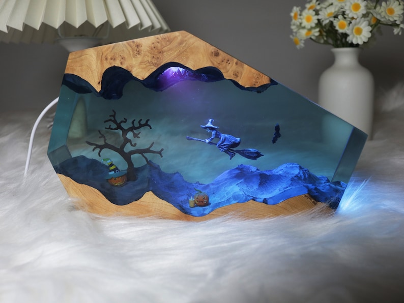May include: A wooden base with a clear resin insert featuring a witch flying on a broomstick, a tree, and a pumpkin. The resin is illuminated with blue light.