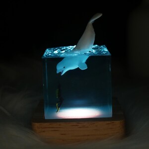 May include: A white beluga whale figurine is suspended in a clear resin cube with a blue tint. The cube is lit from below, casting a soft glow on the whale and the base. A small black and yellow figurine of a diver is at the bottom of the cube.