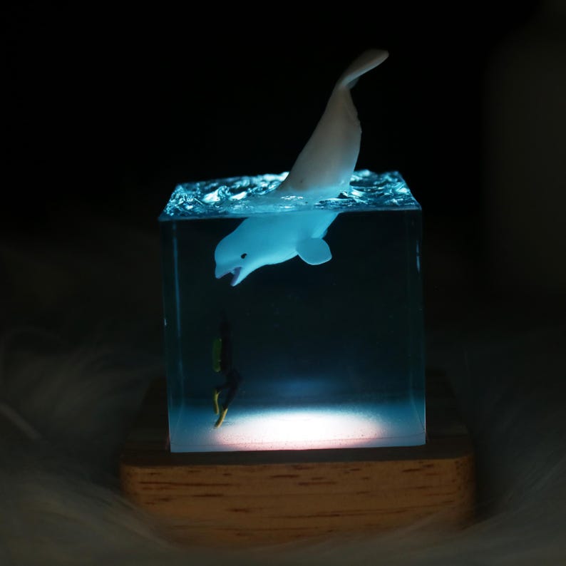 May include: A white beluga whale figurine is suspended in a clear resin cube with a blue tint. The cube is lit from below, casting a soft glow on the whale and the base. A small black and yellow figurine of a diver is at the bottom of the cube.